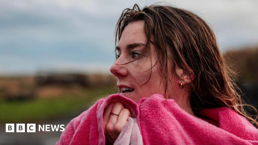 The image shows a close‑up outdoor portrait near the coast of a person with wet hair, wrapped in a bright pink towel and holding it up around their neck. The background is softly blurred, showing a muted coastal landscape with grass, dunes and an overcast sky.
