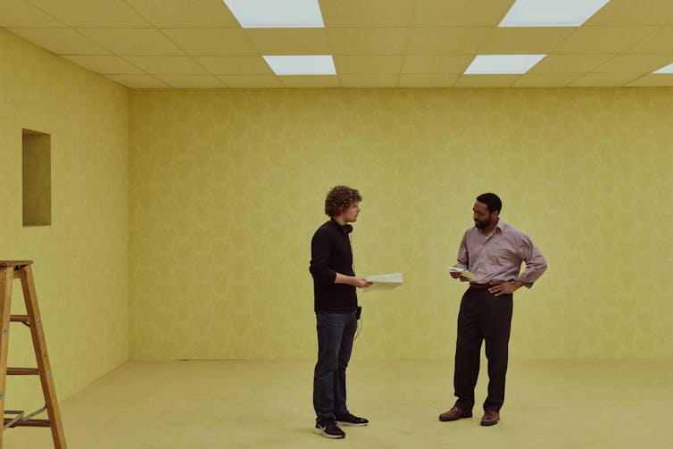 A young director speaks to an actor on a blank, beige-coloured set.