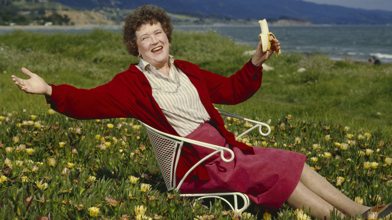 Chef Julia Child smiling and eating a banana in a flowery field with an ocean view