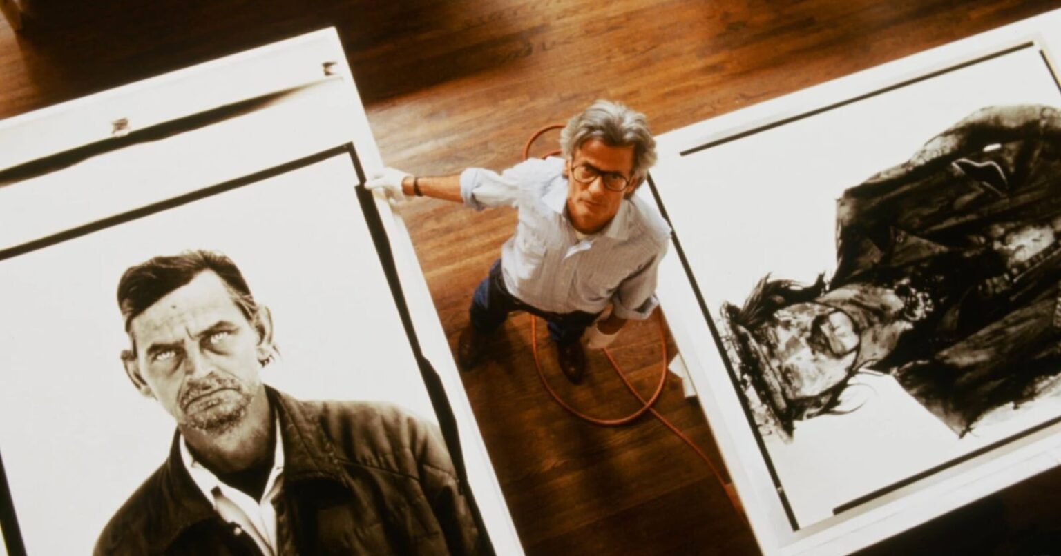 Ron Howard’s Richard Avedon Documentary to Premiere at Cannes Film Festival A man with glasses stands between two large black-and-white portrait artworks on the floor, looking up at the camera. The floor is wooden, and an orange cord is visible beside him.