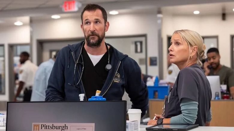 Noah Wyle as Dr. Robby Robinavitch, wearing a navy sweatshirt over black scrubs and a stethoscope around his neck, looking off camera and standing next to Katherine LaNasa as Dana Evans, wearing grey scrubs and a light teal long-sleeved shirt underneath, with her elbow on the counter in The Pitt