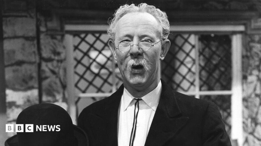 Mr Pastry: British television’s forgotten star A black-and-white photo of a man pictured from the chest up. He is in his 50s, but made-up to look older, with white hair and a drooping white moustache. He is wearing a black jacket and a white shirt, glasses, and is holding a black bowler hat in his right hand, visible at the bottom of frame as he holds it up in front of him. He is standing in front of a stone-walled house with a leaded, diamond-pattern window, with a framed painting visible on the wall through the window, and he is smiling at the camera.