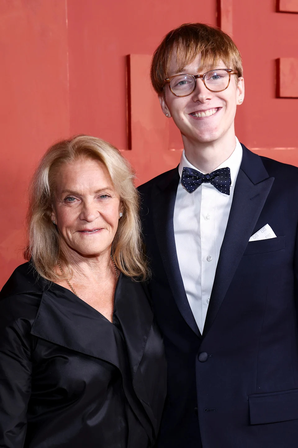 Two people in formal attire smile at an event; one wears a bow tie and glasses, the other a black outfit