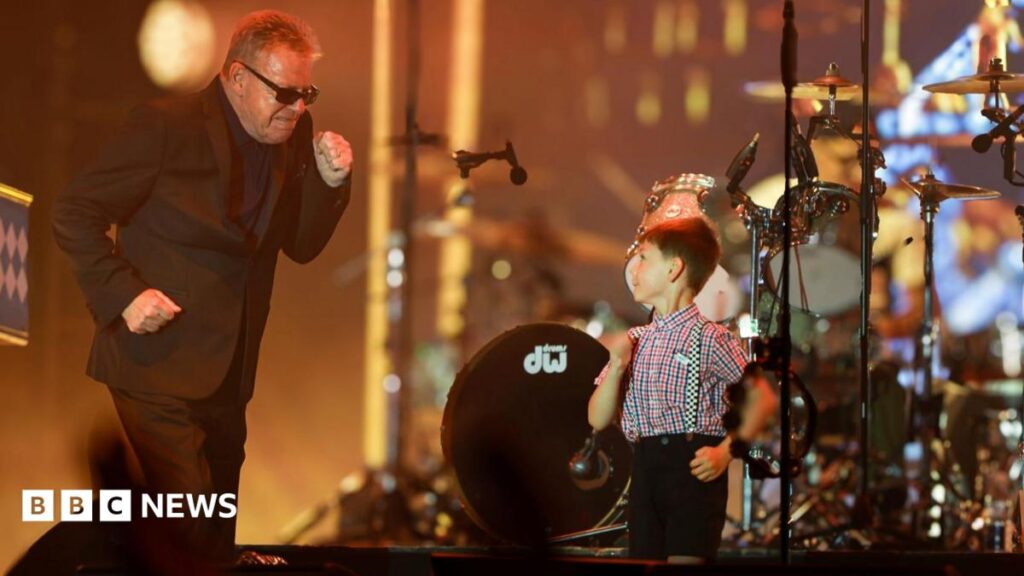 Suggs from Madness with short hair and wearing a dark suit and sunglasses dances with Henry who has brown hair and is wearing black shorts and braces and a patterned shirt