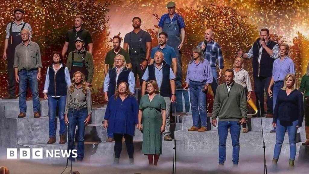 Hawkstone Farmers Choir say mental health message ‘most important’ A large group of performers stands on tiered platforms on a brightly lit stage, appearing to sing together as a choir. A conductor stands at the front, guiding the performance. In front of the stage, a panel of judges sits at a table, watching. The background features warm, glowing lights and foliage-like scenery, while an audience in the foreground raises hands and watches the show.