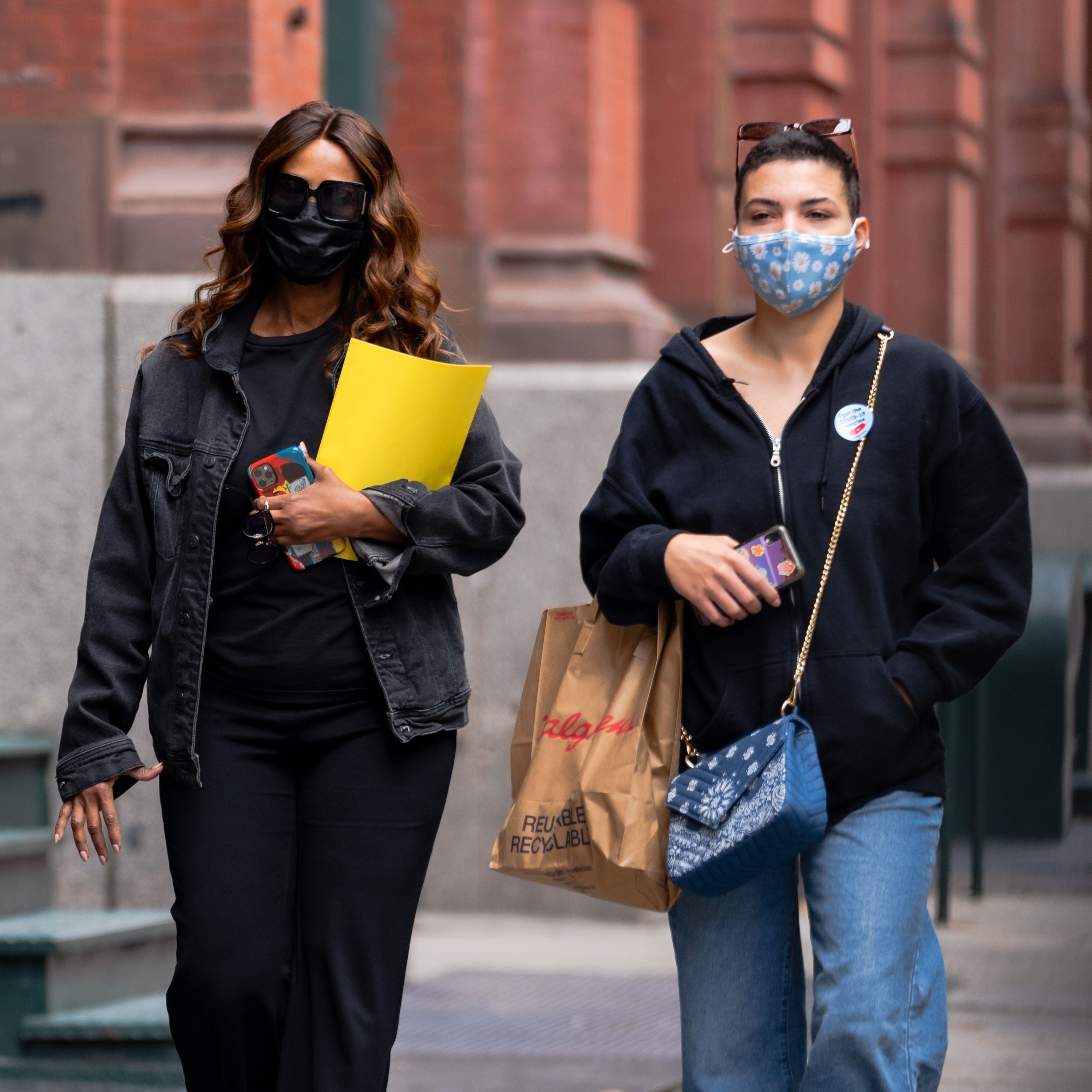 Two people walking on a street, wearing face masks and casual outfits. One holds a yellow folder; the other carries a shopping bag