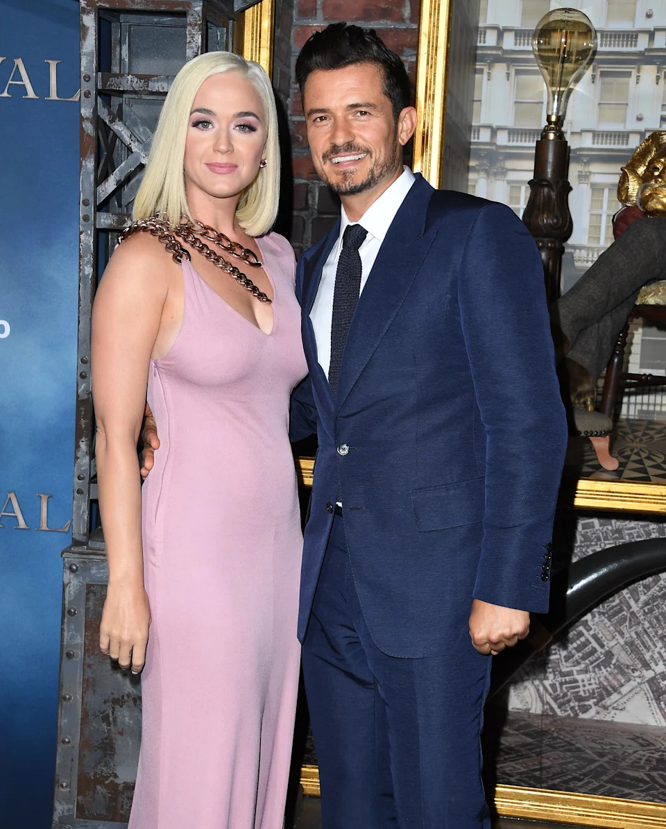 Two people pose on a red carpet; one in a sleek gown with chain detail, the other in a tailored suit