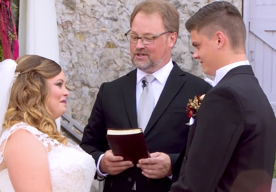 A couple stands facing each other at an outdoor wedding ceremony, officiated by a man holding a book