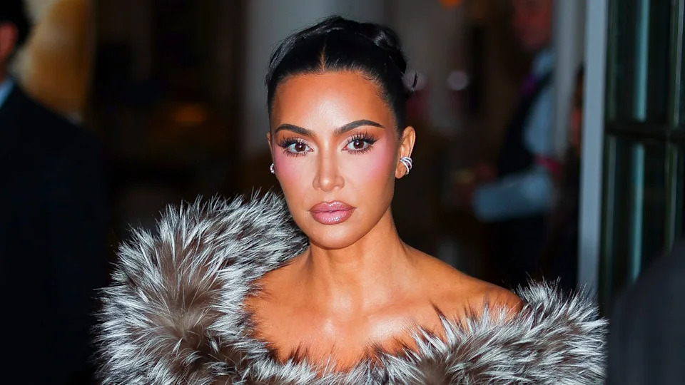 Kim Kardashian wearing a fur outfit, with her hair styled up, and makeup highlighting her features, looking directly at the camera.