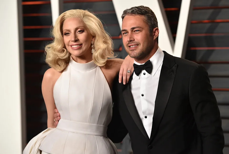 Two individuals on the red carpet. One is in an elegant, sleeveless gown with dramatic styling; the other in a classic black tuxedo