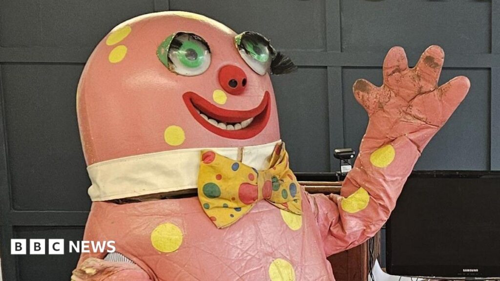 Mr Blobby costume sold at auction in Bristol for £8,500 A large, pink Mr Blobby costume is pictured with big googly eyes, a yellow bow tie and dots. It is raising its arm up with its hands out, waving.