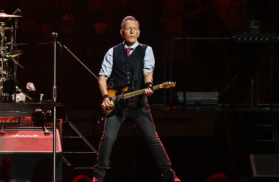 Musician performing energetically on stage with a guitar, wearing a vest, tie, and jeans. Stage lights illuminate the scene