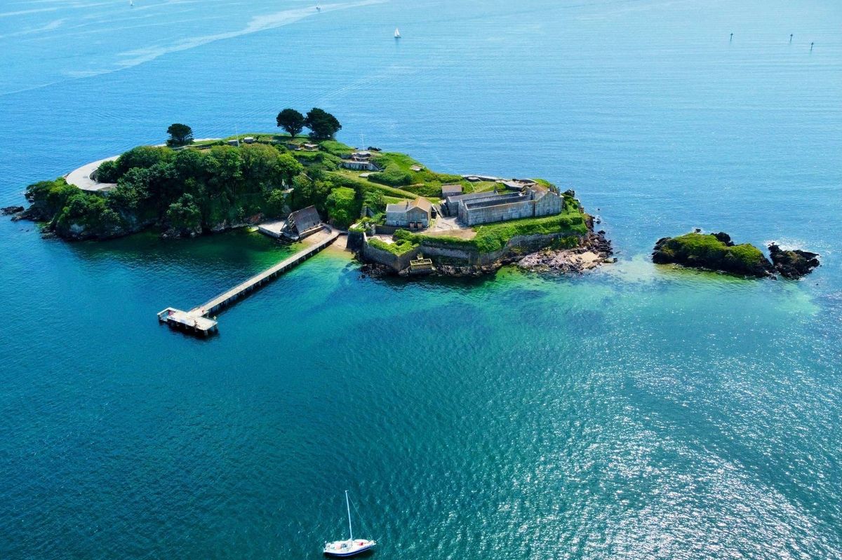 An aerial view of a small island with a dock extending into the water, surrounded by a serene blue ocean. The island features lush greenery and a structure amidst the trees, creating a picturesque setting.