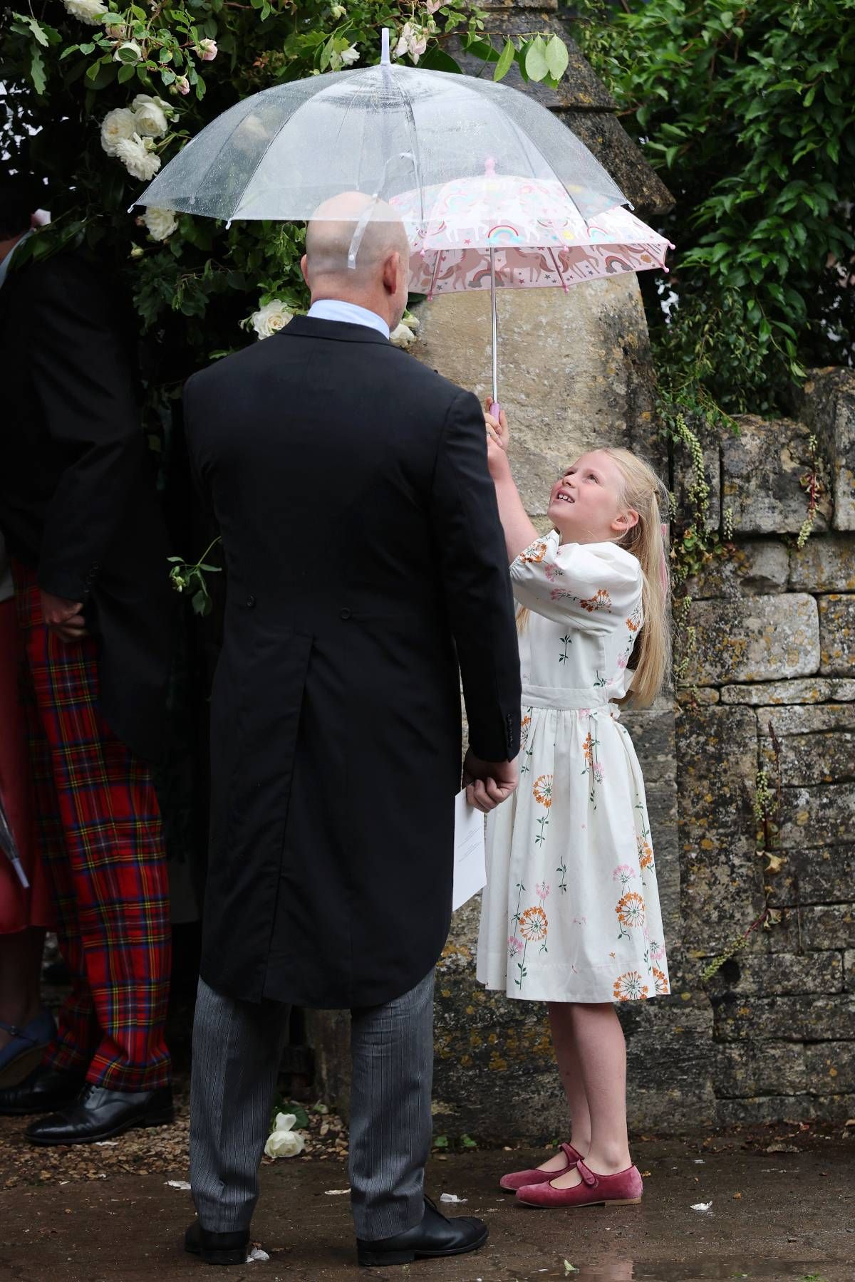 A young girl in a white dress reaches up towards an individual dressed formally in a black suit, standing against a backdrop of a stone wall with foliage.