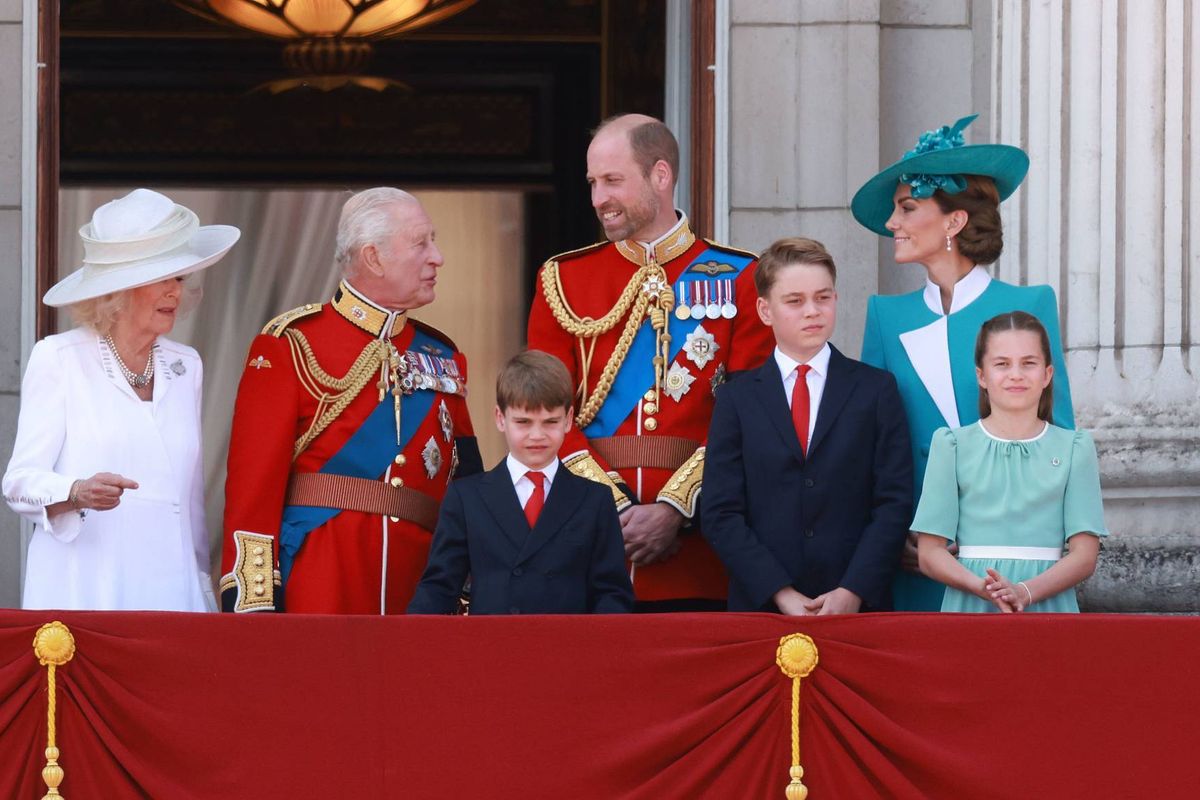 A group of individuals, possibly members of a royal family, are gathered on a balcony adorned with red drapery. They are dressed in formal attire, with some wearing hats and military uniforms, and they are facing the camera, possibly attending an official event.