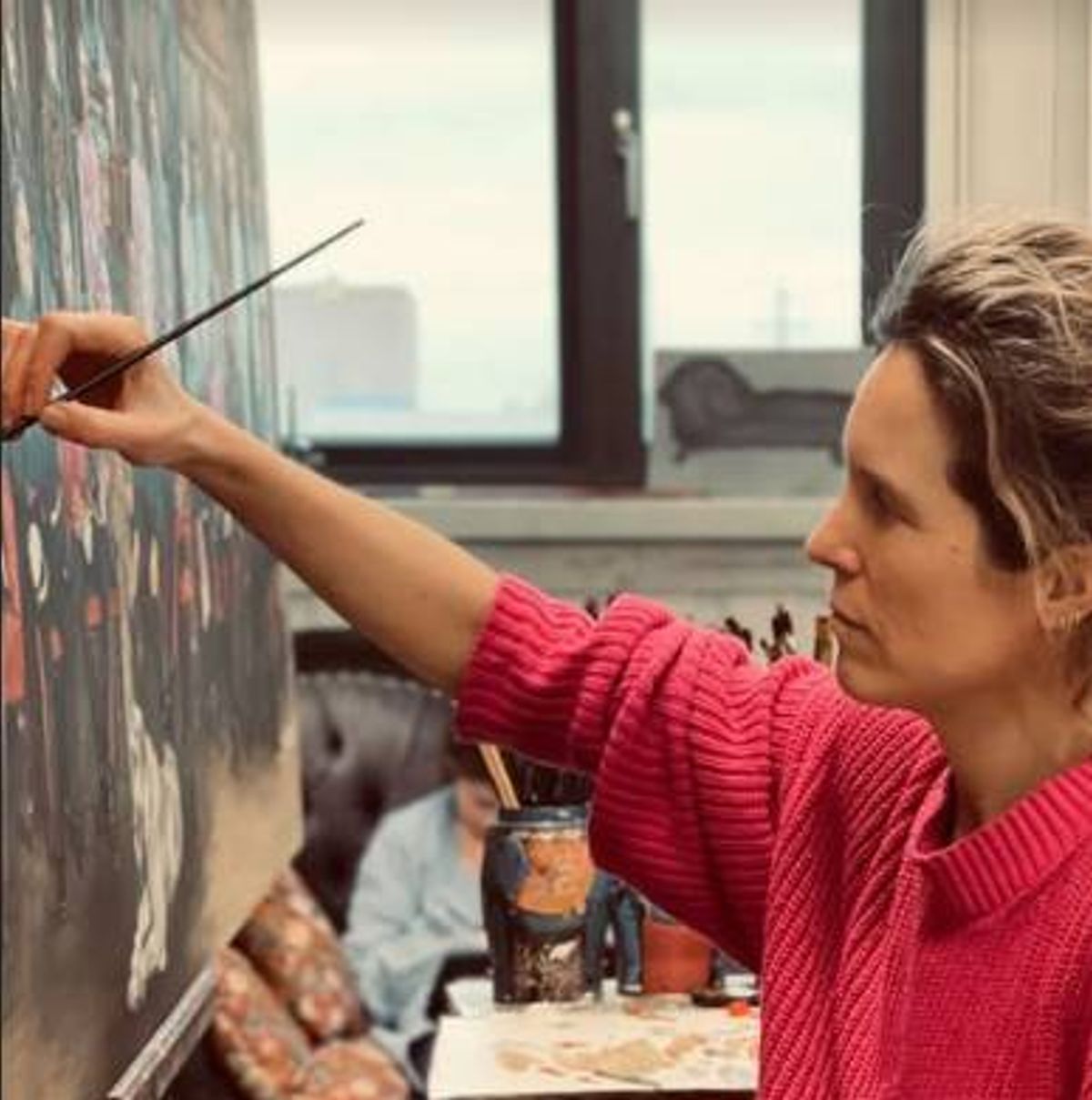 An individual, dressed in a red long-sleeved garment, is engaged in the act of painting on a large canvas, with a blurred figure seated at a table in the background.