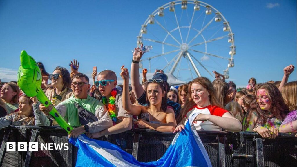 A crowd of people wearing summer clothes stand behind a barricade watching a performance. Two people at the front hold a Scotland flag. There is a ferris wheel behind them.