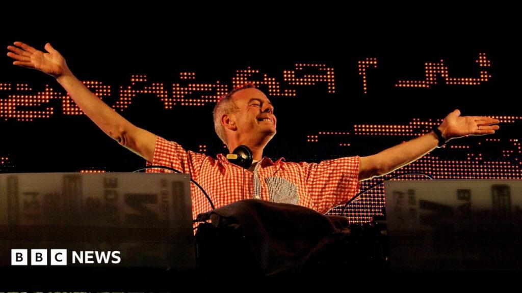 A man wearing a red and white checked shirt, with headphones draped around his neck, stands behind a DJ booth, arms outstretched.