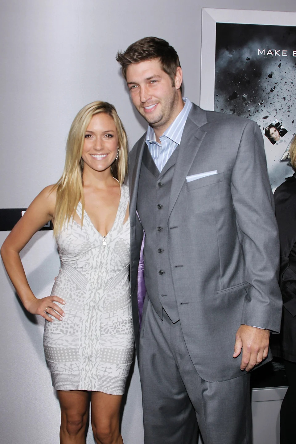 Two individuals pose on a red carpet; one wears a tailored grey suit, and the other wears a sleeveless patterned dress