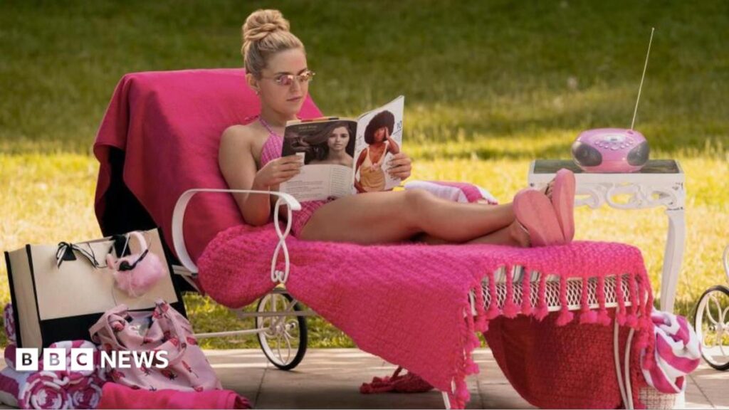 A woman in a pink bikini lies on a deck chair covered in pink blankets, reads a magazine. there are pink towels, a tote bag and a radio next to her.