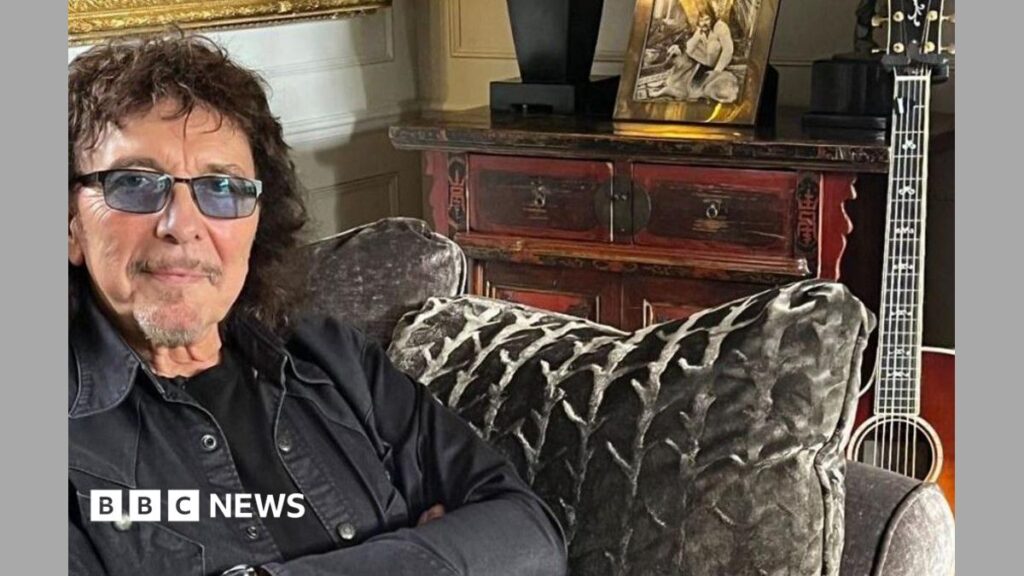 Black Sabbath’s Tony Iommi made an MBE in King’s Birthday Honours A man wearing tinted spectacles and a dark jacket sits on a sofa, with a guitar propped up against it