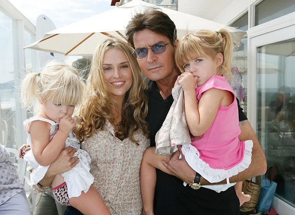 Two adults pose with two young children outdoors, near a patio with umbrellas, enjoying a sunny day