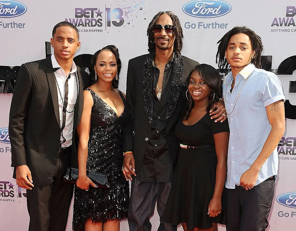 Group of five people at a red carpet event, elegantly dressed. The person in the center wears a black jacket with ornate detailing