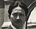 A black and white photo of Kiyoshi Tanimoto in front of damaged buildings.