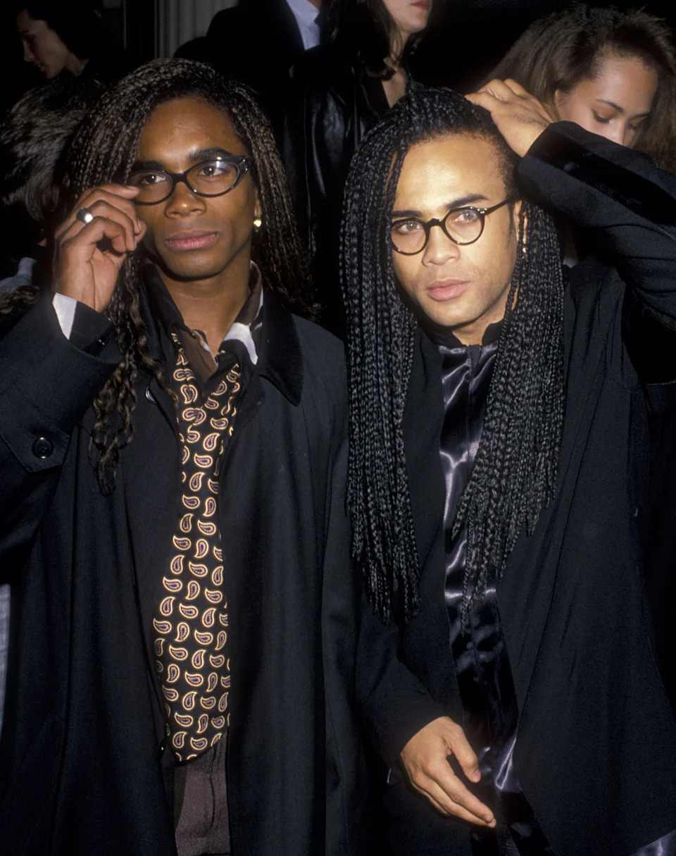 Milli Vanilla with long braided hair wearing glasses and stylish coats at an event