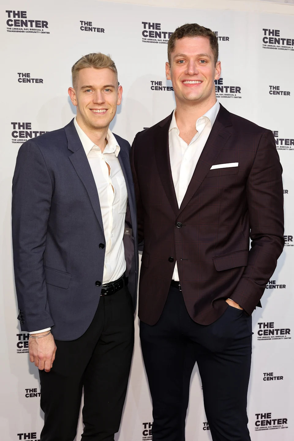 Two people in suits, one in a dark blazer and the other in a darker suit, smile on the red carpet at a formal event