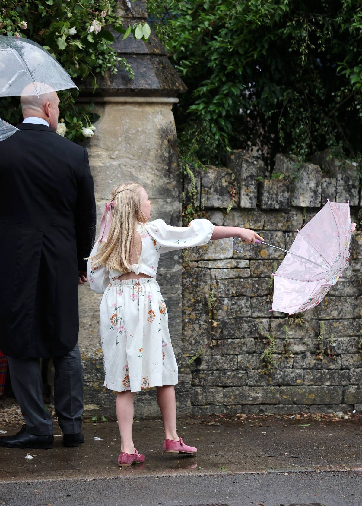 An individual dressed in a black suit stands next to a child in a white dress, both holding pink umbrellas, positioned in front of a stone wall adorned with greenery.