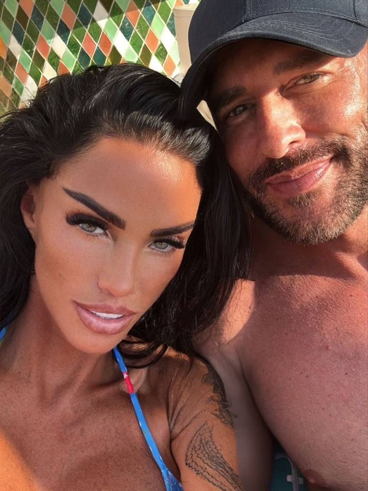 An individual with dark hair and prominent facial features is posing closely with another individual, who is shirtless and sporting a beard. Both individuals are smiling and appear to be in a relaxed, possibly romantic setting.