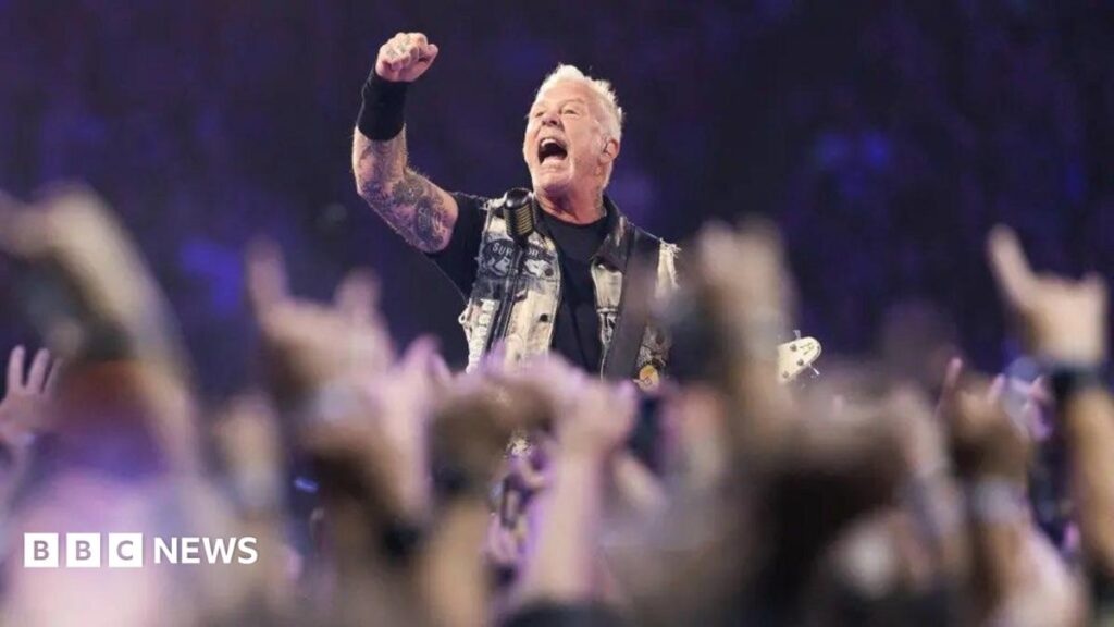 Metallica front man, James Hetfield, punches the air wearing a waistcoat, black T-shirt and wrist sweat band with short blonde hair