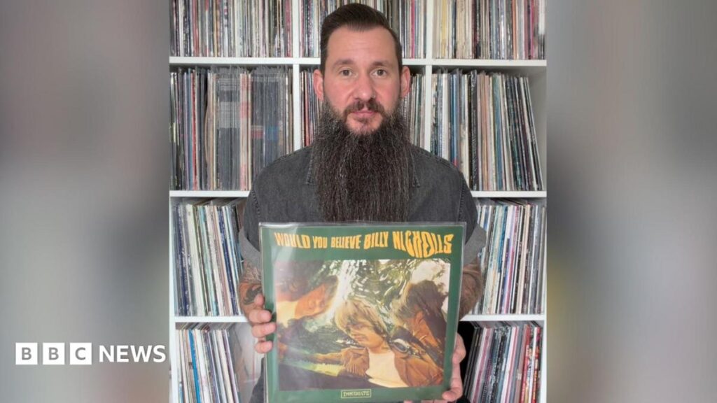 Martin is holding Billy Nicholls record, would you believe.  The cover is green with yellow text.  The image is a wavy effect photo of the main artists involved. Martin has dark hair and a very long beard. He is sat infant of loads of vinyl records in shelves