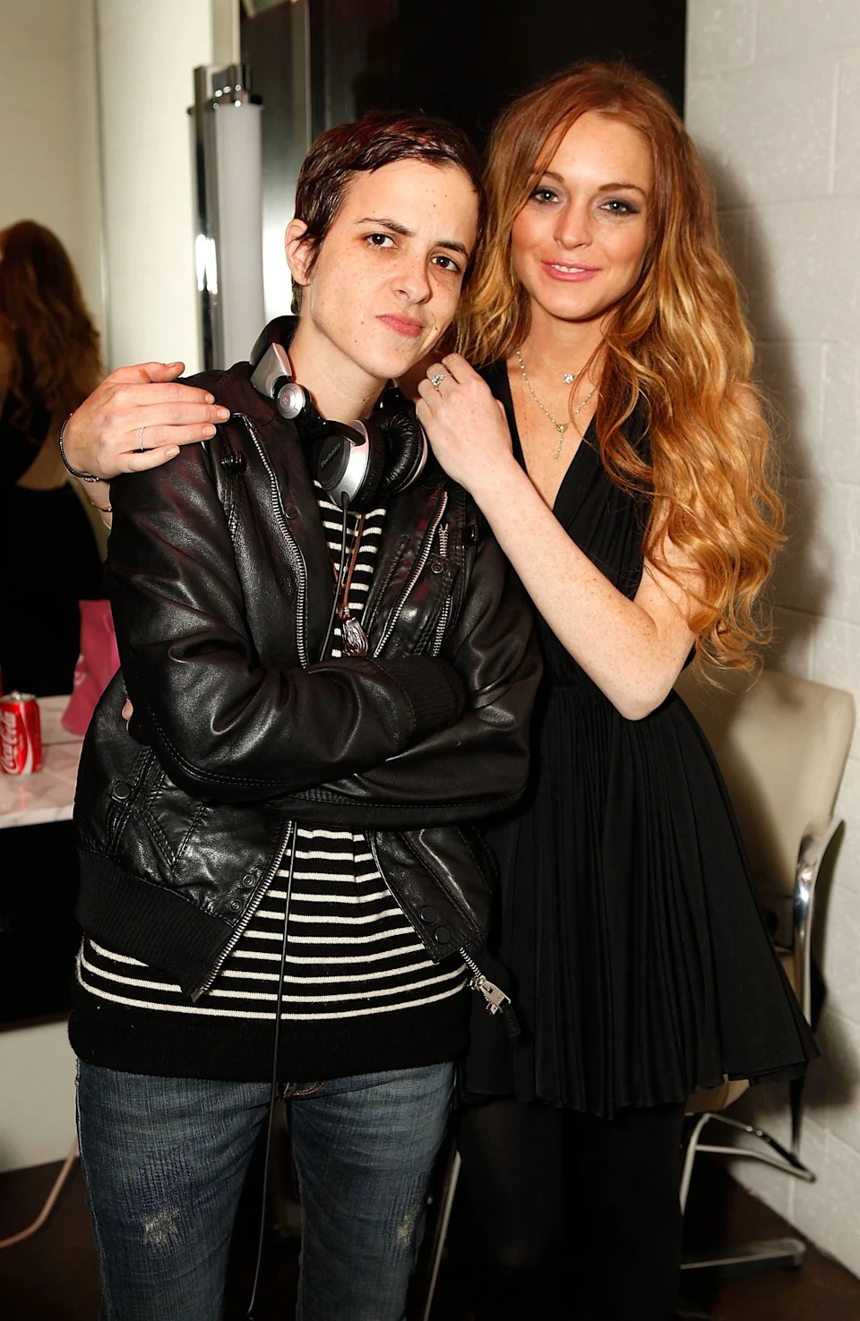 Two individuals pose together; one in a leather jacket with headphones, the other in a sleeveless dress. They're at an event with a friendly vibe