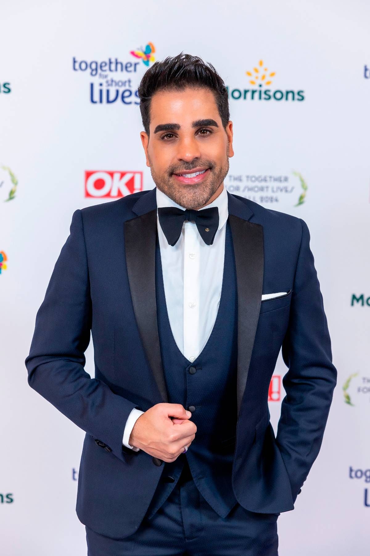 An individual dressed in a formal suit and bow tie, standing against a backdrop featuring various logos and text, including "OK!" and "Morrisons", with a cheerful expression.