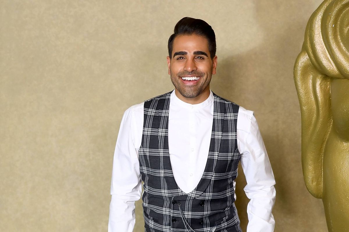 An individual stands smiling, wearing a white t-shirt and a black and white checkered vest against a beige backdrop.