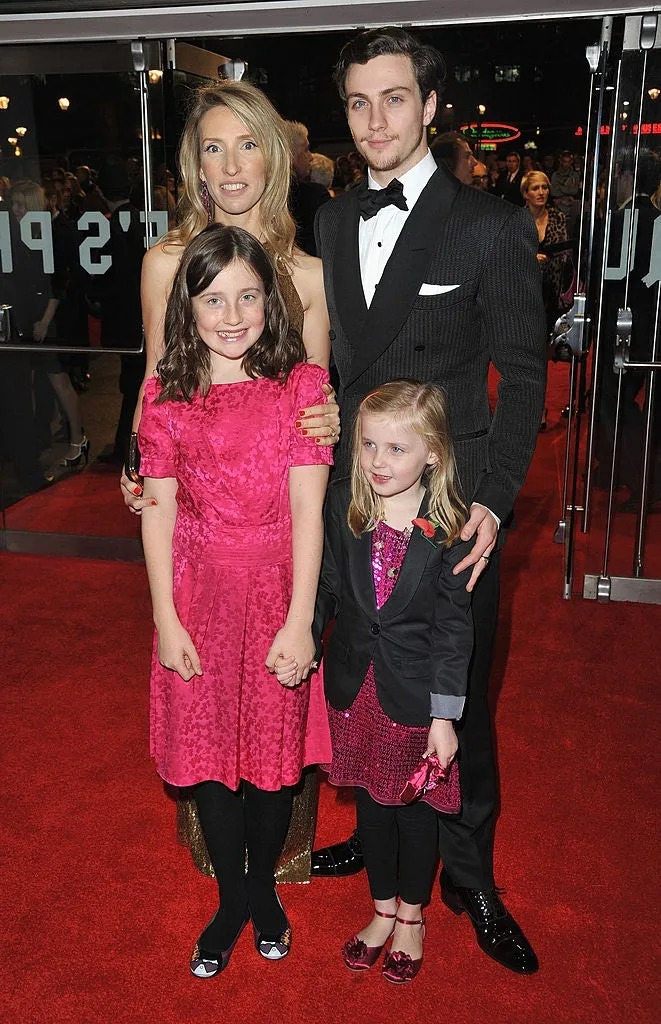 Two adults and two children pose on a red carpet. The adults are in formal attire, the man in a suit and the woman in a strapless dress