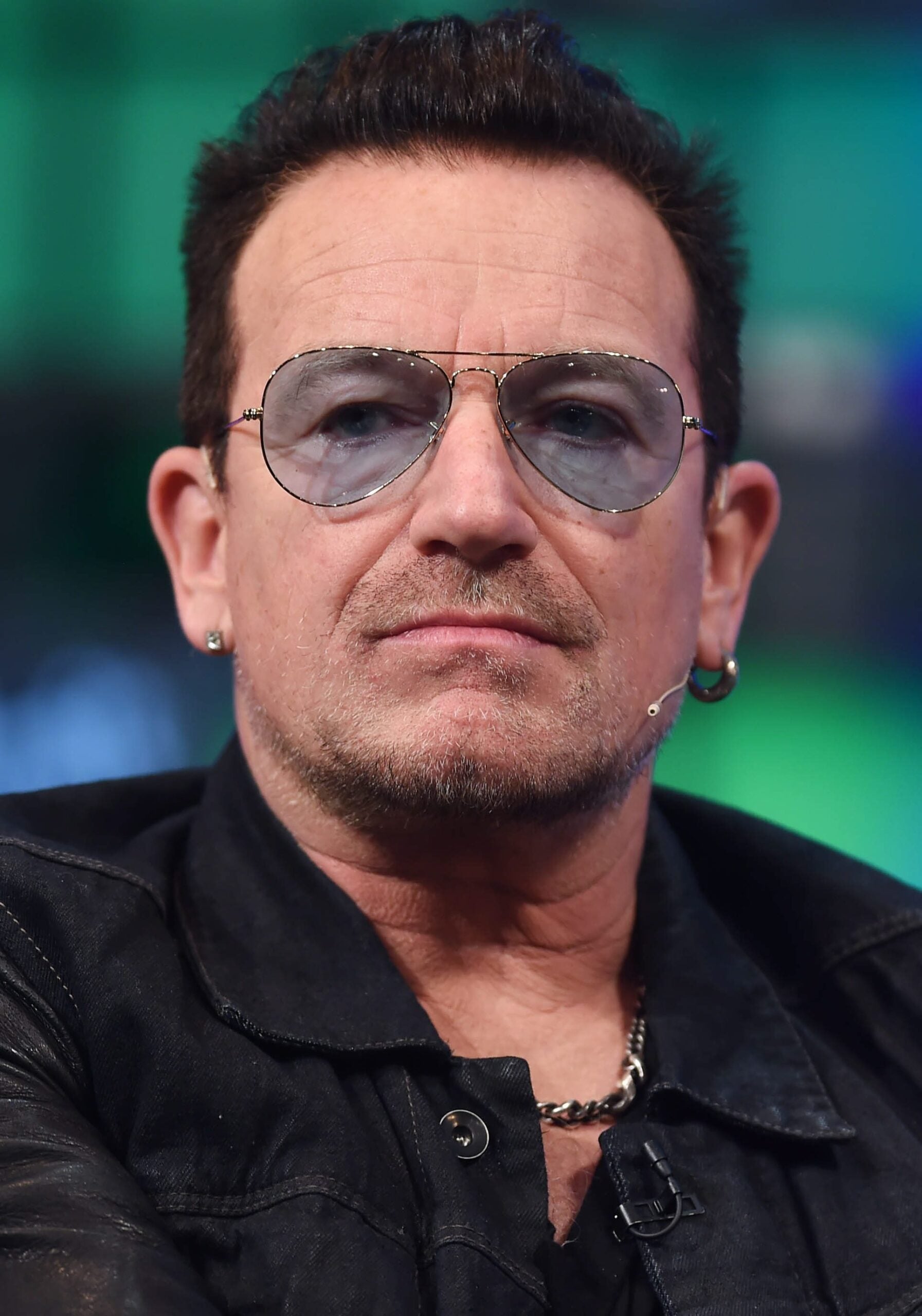 A man with short dark hair, wearing aviator sunglasses, a black jacket, a necklace, and small hoop earrings, looks directly at the camera against a blurred background.