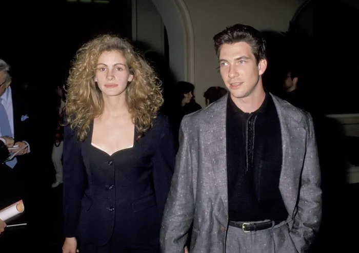 Two individuals at an event; one wears a structured blazer and has curly hair, the other wears a suit jacket over a dark shirt