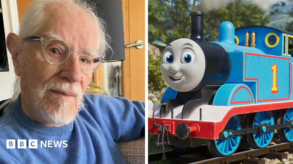 A blue train engine with a grey smiling face. It has a yellow number one on the side of it, and the track it is running along is surrounded by countryside.