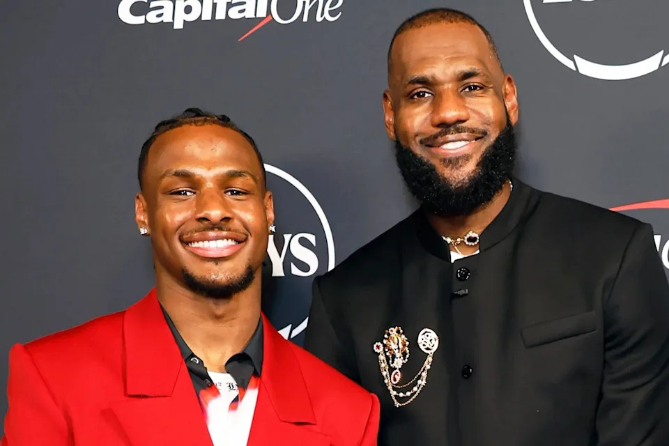 Two individuals at a formal event; one in a red jacket and the other in a black suit with decorative pins. They are smiling at the camera