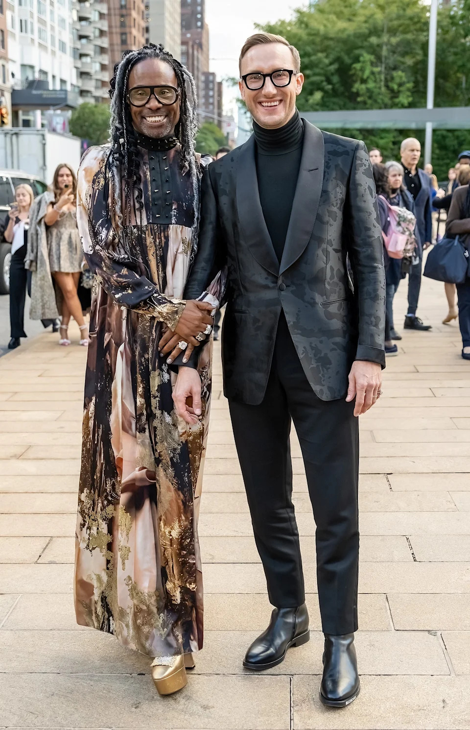 Two people stand on a city street. One wears a long patterned gown; the other wears a sleek suit with a turtleneck