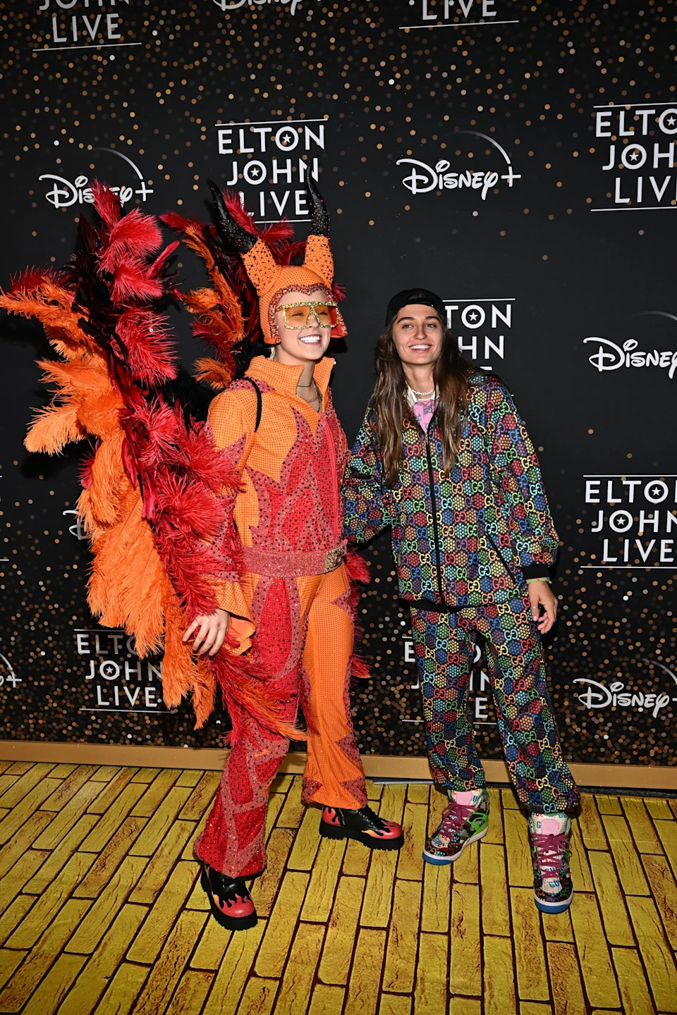 Two individuals pose on the red carpet at an Elton John event. One wears a vibrant, feathery costume, and the other sports a colorful, patterned outfit