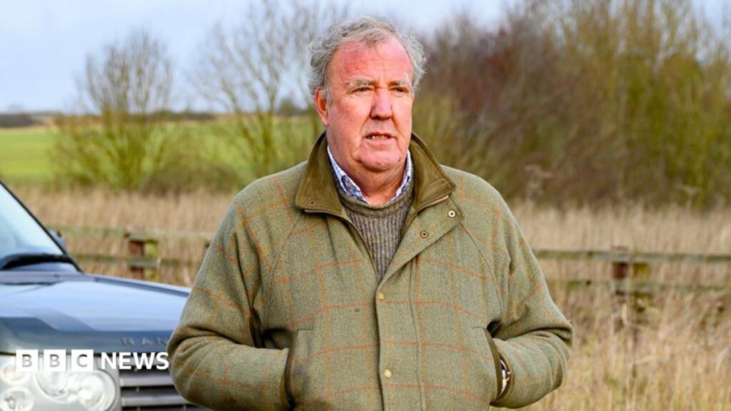 Jeremy Clarkson's health dilemma 'common among farmers' – BBC Jeremy Clarkson is pictured standing in front of a Range Rover car looking into the distance. He is wearing a green tweed coat and he has his hands in the pockets of the coat. There is a fence behind him and beyond that is a green field and some trees.