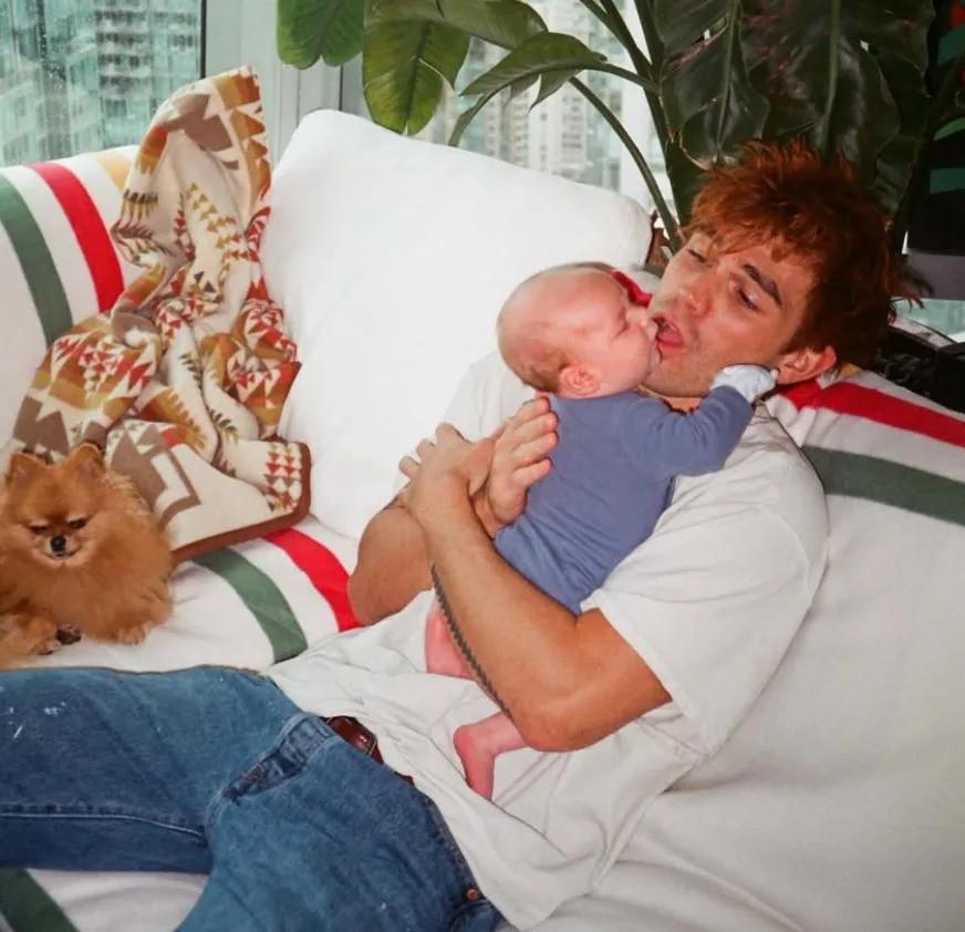Person in casual clothes holding a baby, sitting on a sofa with a small dog nearby, in a cozy setting