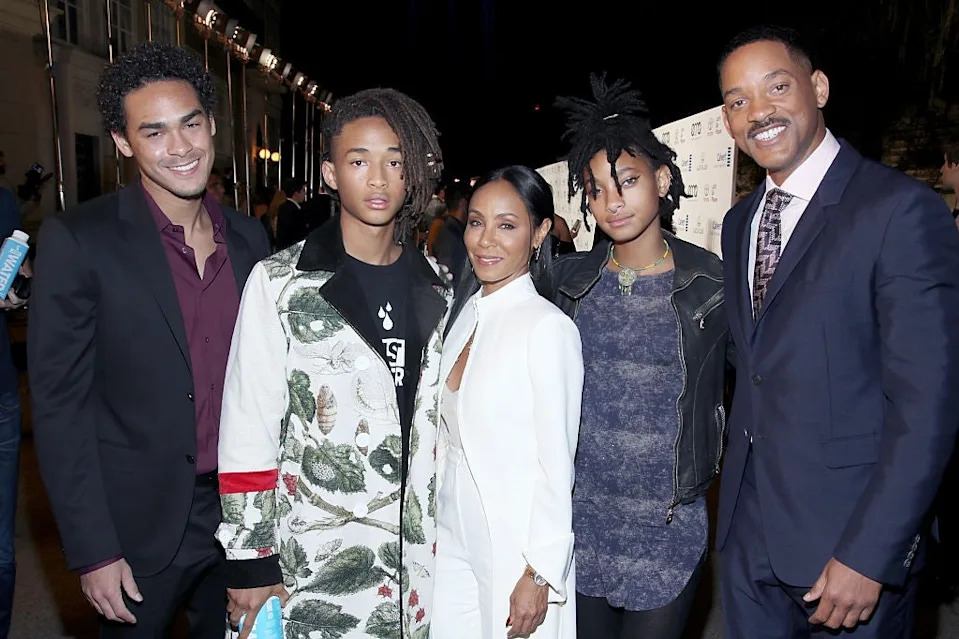 Five people pose together on a red carpet; some wear stylish coats and casual outfits