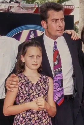 A man in a suit with a patterned tie stands with a young girl in a floral dress at a public event