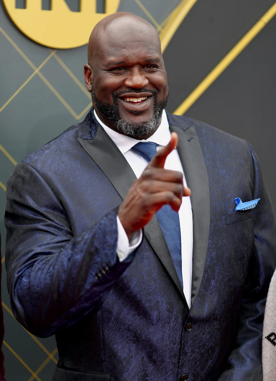 Person in a dark suit with a tie, smiling and pointing on a red carpet