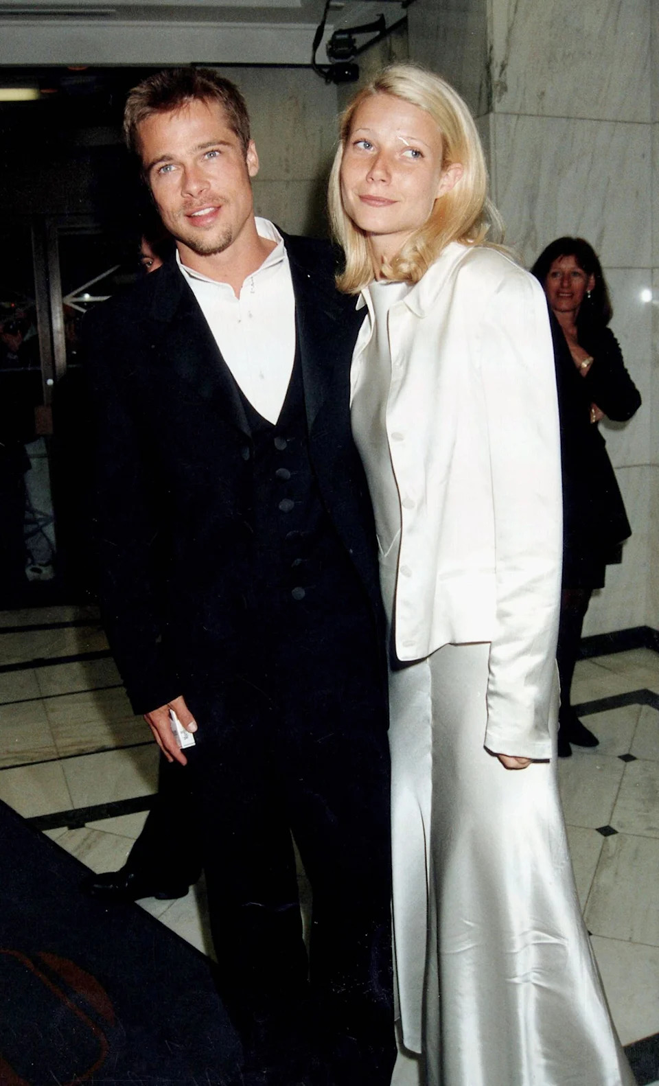 Man in a suit and woman in a satin jacket and skirt at a formal event, posing for a photo on a marble floor